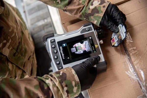A close up shot of a person in camo and nitrile gloves holding a Rigaku ResQ CQL device above a cardboard box, using the device's camera function to scan a tag.